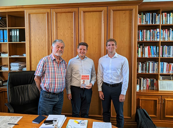 Three men standing in front of a bookcase. One of the men is holding a book