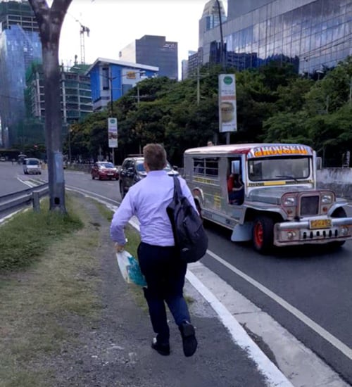 Man running next to a busy road. He is carrying a backbag and a plastic bag