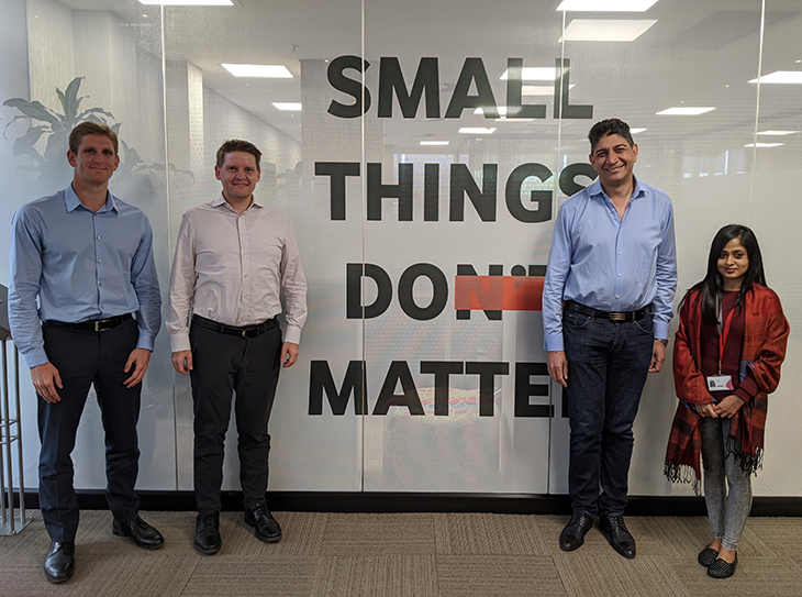 Four people standing in front of a glass wall that has "small things do matter" written on it